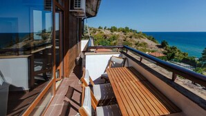 A balcony with a wooden table and chairs, overlooking a coastal landscape with a clear view of the sea and hills.