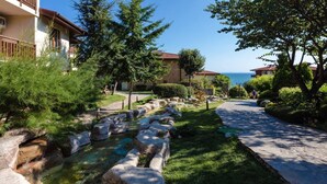 A landscaped pathway with a small stream, surrounded by greenery and buildings with a view of the sea.