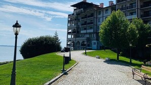 A cobblestone pathway leading to a multi-story building with balconies, a lamp post, and a view of the sea.
