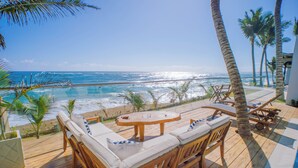 A wooden deck with a round table and cushioned seating facing a beach and ocean.