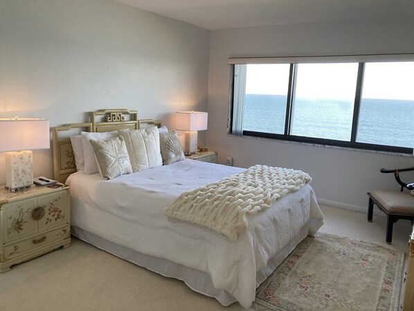 A bedroom with a large bed, two bedside tables with lamps, a chair, and a view of the sea through a window.