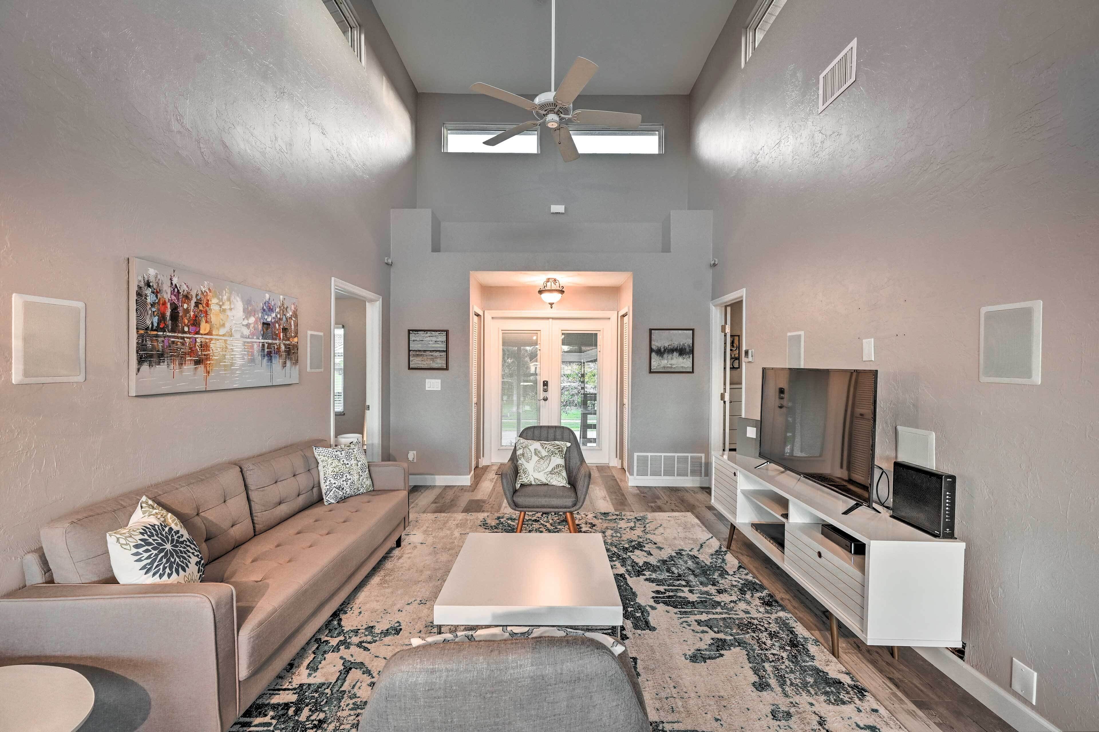 A spacious living room with a large sofa, a coffee table, and a TV on a white entertainment unit. There is a ceiling fan, a large painting on the wall, and a rug on the floor.