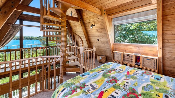 A wooden cabin with a spiral staircase, a large bed with a colorful bedspread, and a view of the sea through large windows.