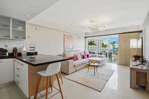 Una cocina moderna con una isla central, una zona de comedor con sofá y sillas, una sala de estar con un ventanal, y un balcón con vistas a edificios y palmeras.