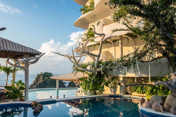 Un área junto a la piscina con vista despejada al mar, una pérgola y un edificio de varios niveles con vegetación.