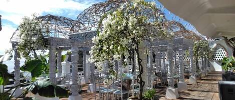 Un espacio al aire libre cubierto con columnas y arcos blancos, plantas en macetas y una terraza de madera.