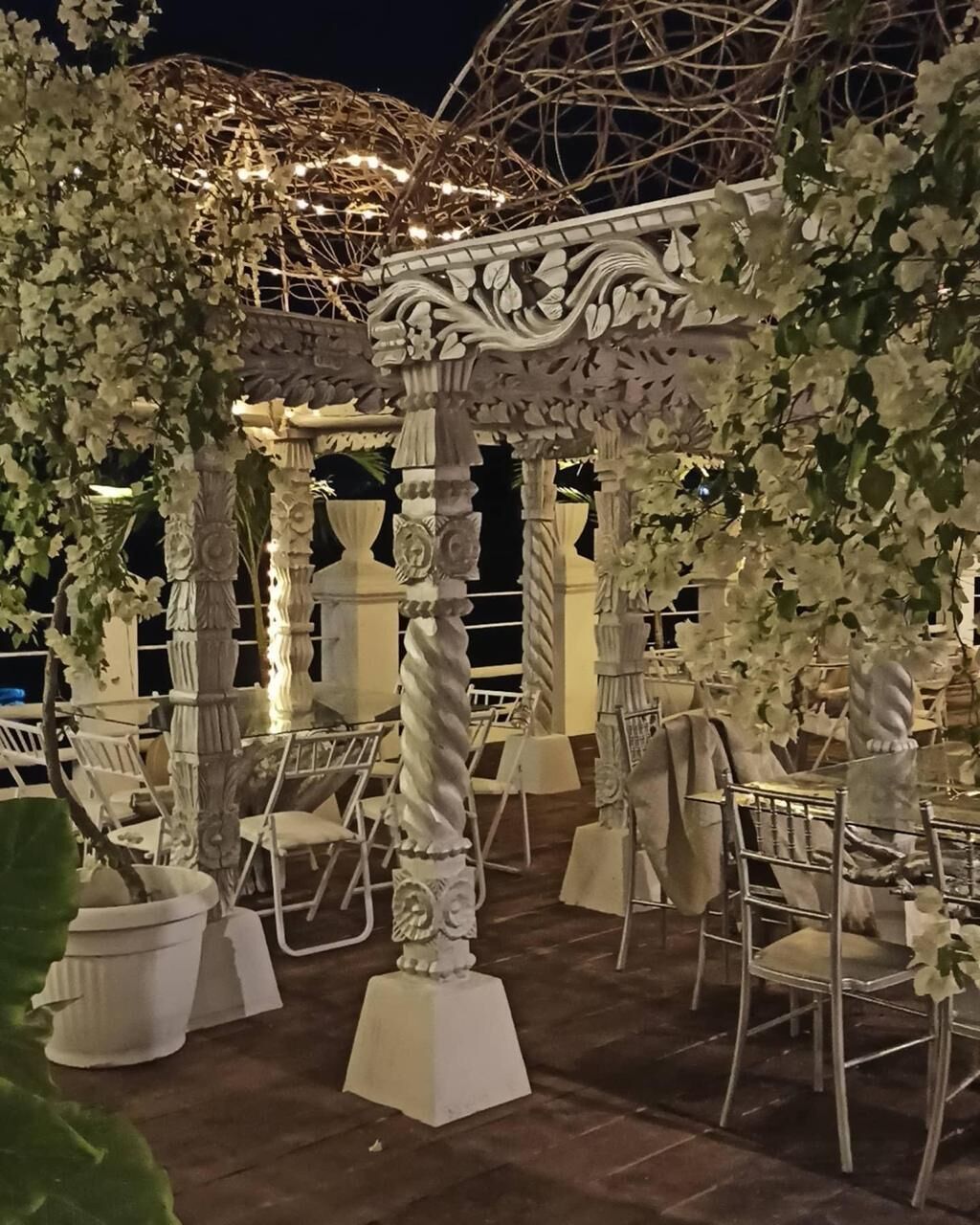 Un área de comedor al aire libre con sillas y mesas blancas, columnas blancas ornamentadas y decoraciones florales.