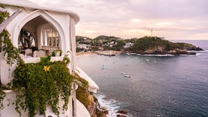 A coastal hotel with a white facade, a balcony with chairs, and a view of the sea and a small island.