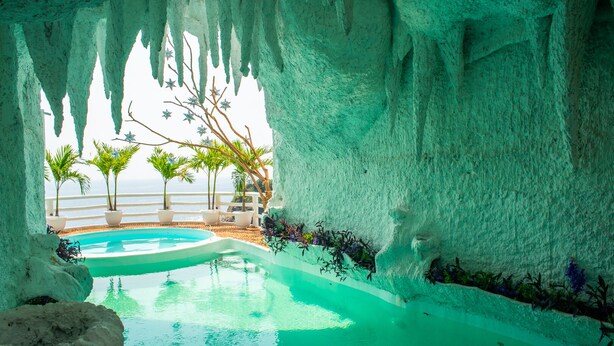 A pool with a view of the ocean, surrounded by white rock walls and palm trees.