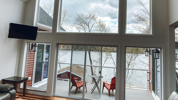 Une pièce dotée de grandes fenêtres donnant sur un lac, d’un téléviseur fixé au mur et d’une terrasse avec des meubles d’extérieur.