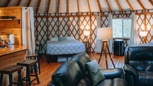 Interieur van een joert met een houten vloer, een bank, een bed, een klein keukenhoekje en een plafondventilator.
