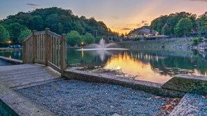 A wooden dock leading to a calm lake with a fountain, surrounded by trees and a building in the background.
