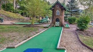 A miniature golf course with a treehouse structure, green artificial turf, and a small pond.