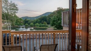 A balcony with two wicker chairs, a wooden railing, and a view of a lake with trees and hills in the background.