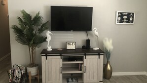 Uma sala de estar com televisão na parede, um armário branco com o símbolo Wi-Fi, uma planta em vaso e um enfeite de cavalo-marinho.
