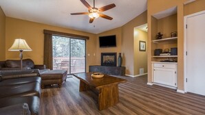 Un salon comprenant un canapé, un pouf, une table basse en bois, un ventilateur de plafond, un téléviseur, une cheminée et une vue à travers la porte vitrée.