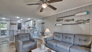 A modern living room with a grey sofa, a coffee table, and a ceiling fan. There is a kitchen area in the background with white cabinets and a dining table. - Keowee Key ~ Cozy Condo ~ Golf Course View - Amenities (Salem)