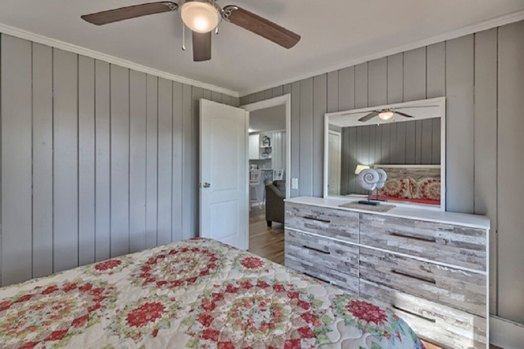 Une chambre à coucher comprenant un lit, une commode, un ventilateur de plafond et une porte donnant sur un couloir.