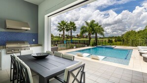 Eine moderne Außenküche mit Grill, Esstisch und Stühlen. Ein Schwimmbad mit Liegestühlen und Blick auf einen Golfplatz und Palmen.