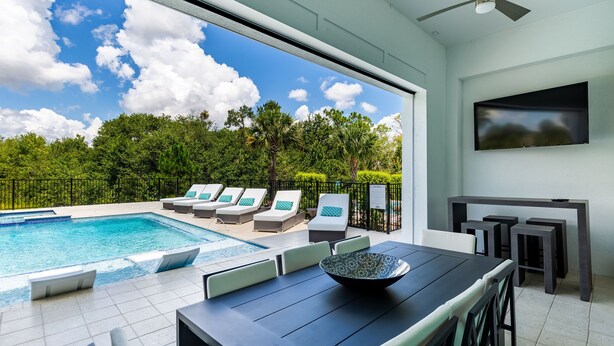 Ein moderner Poolbereich im Freien mit einem Esstisch und Stühlen, einem an der Wand montierten Fernseher und Blick auf Himmel und Bäume.