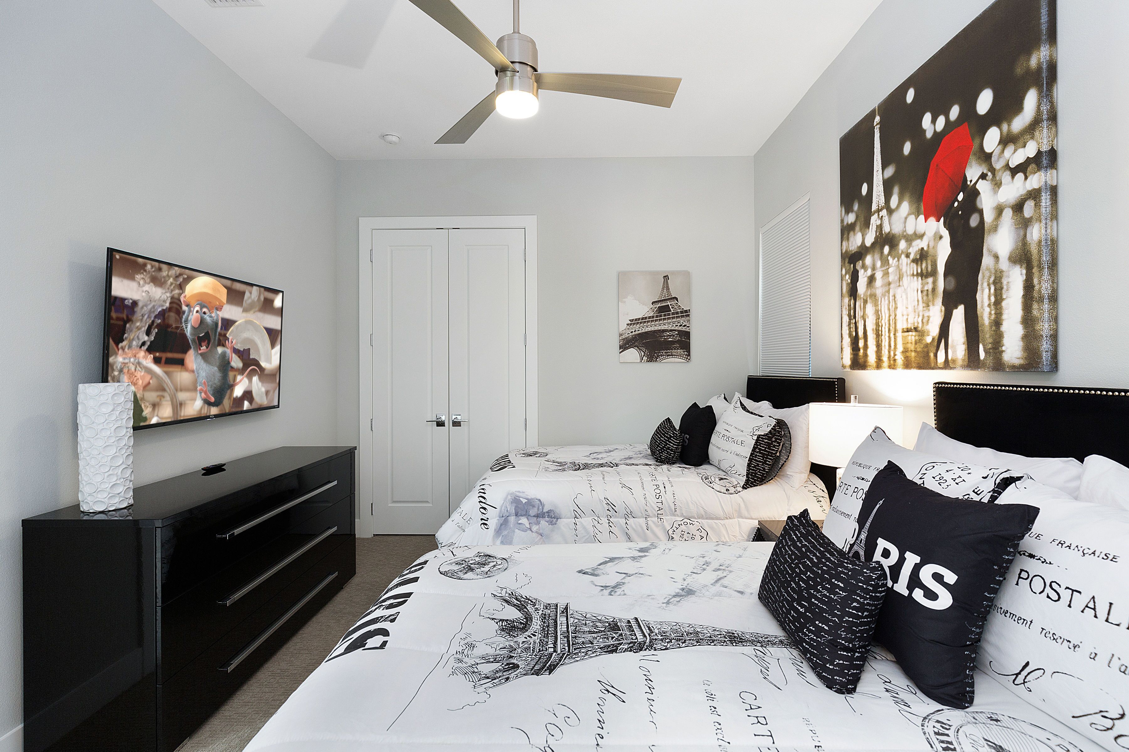 A bedroom with a bed, a TV, a ceiling fan, and artwork on the walls.