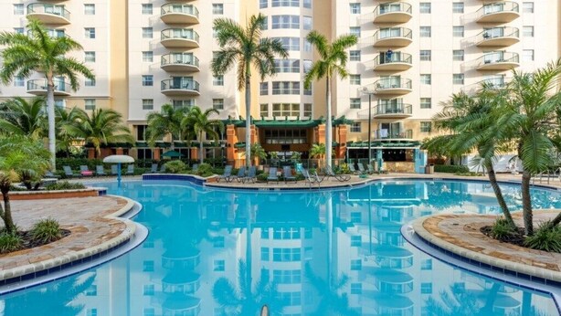 A hotel with a large swimming pool surrounded by palm trees and lounge chairs. - Pompano Beach | Group Golf Vacation | Pool + Golf | 4BR Sleeps 12 (Pompano Beach)