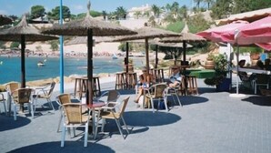 Un café situé en bord de mer, avec une terrasse, des parasols et une vue sur la plage et l’océan.