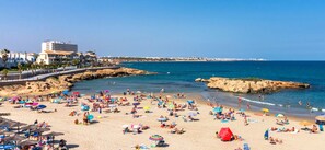 Ein Strand mit badenden Personen, Sonnenschirmen und einer felsigen Küstenlinie.