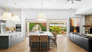 Une cuisine spacieuse dotée d’un îlot central, d’un éclairage moderne suspendu et offrant une vue sur un jardin luxuriant grâce à une arche ouverte.