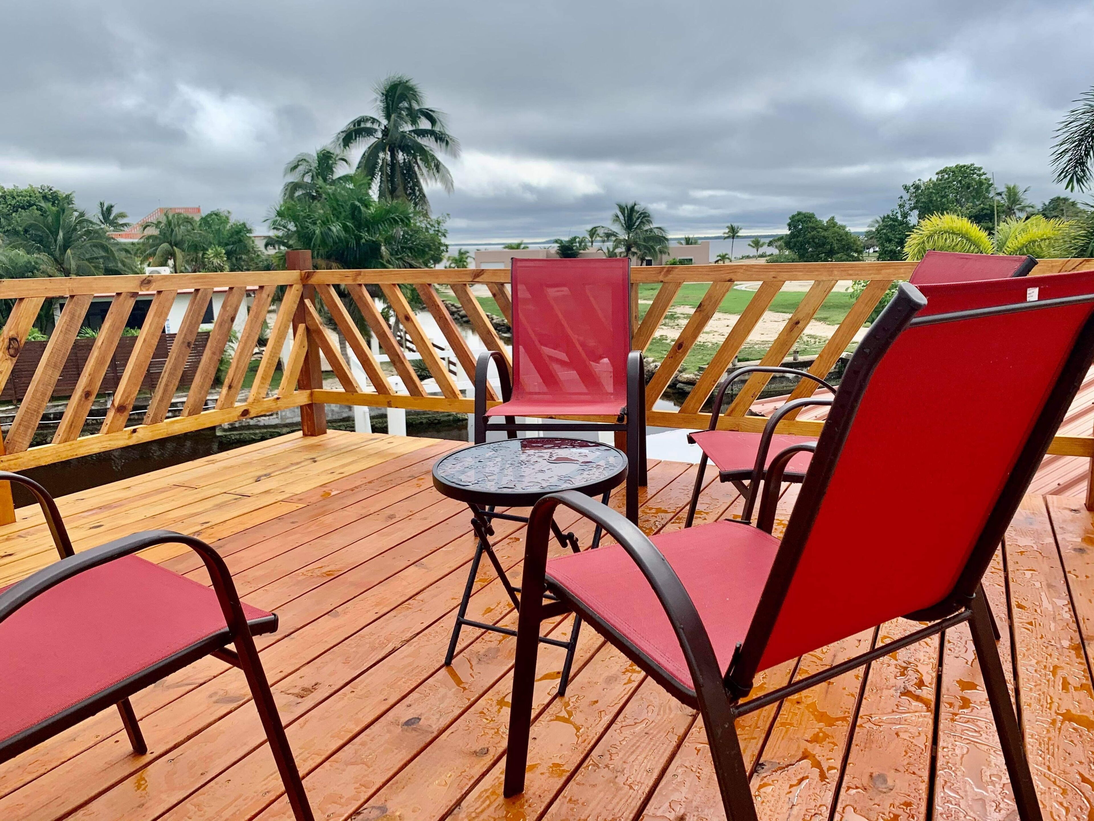 A wooden deck with a table and red chairs, overlooking a beach and palm trees.