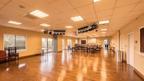 A spacious room with a wooden floor, multiple tables and chairs, ceiling-mounted lighting, and large windows.