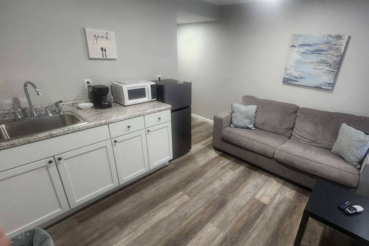 A compact kitchen with white cabinets, a sink, a microwave, and a refrigerator. There's a gray sofa with patterned cushions and a small black table with a remote control.