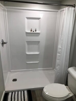 A bathroom with a white shower enclosure, a toilet, and a small shelf with toiletries. - Rockwood Resort Motel (Lake Ozark)