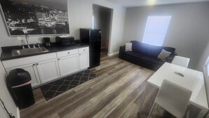 A compact kitchen with white cabinets, a black refrigerator, and a black countertop. There's a black trash bin and a small rug. A white table and a black couch are in the living area, with a window providing natural light. - Rockwood Resort Motel (Lake Ozark)