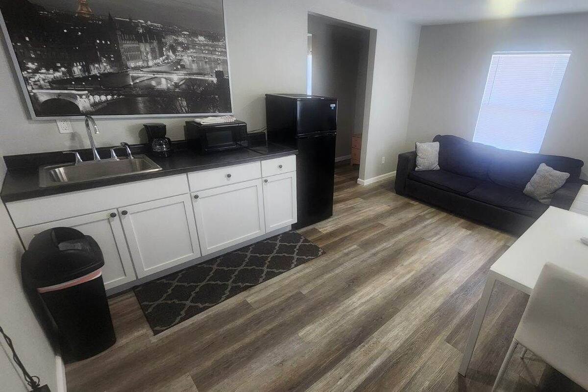 A compact kitchen with white cabinets, a black refrigerator, and a black countertop. There's a black trash bin and a small rug. A white table and a black couch are in the living area, with a window providing natural light.