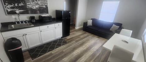 A compact kitchen with white cabinets, a black refrigerator, and a black countertop. There's a black trash bin and a small rug. A white table and a black couch are in the living area, with a window providing natural light.