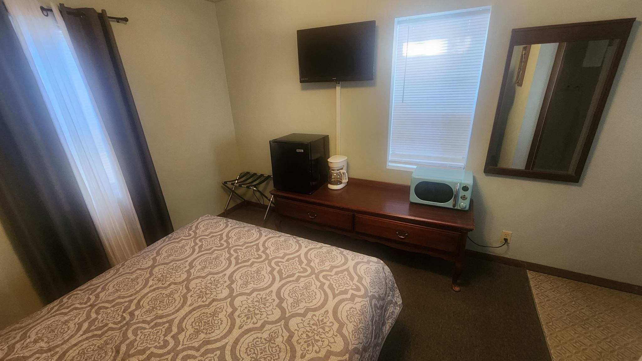 Habitación de hotel con cama, cómoda, cafetera, microondas, televisor y ventana con persianas.