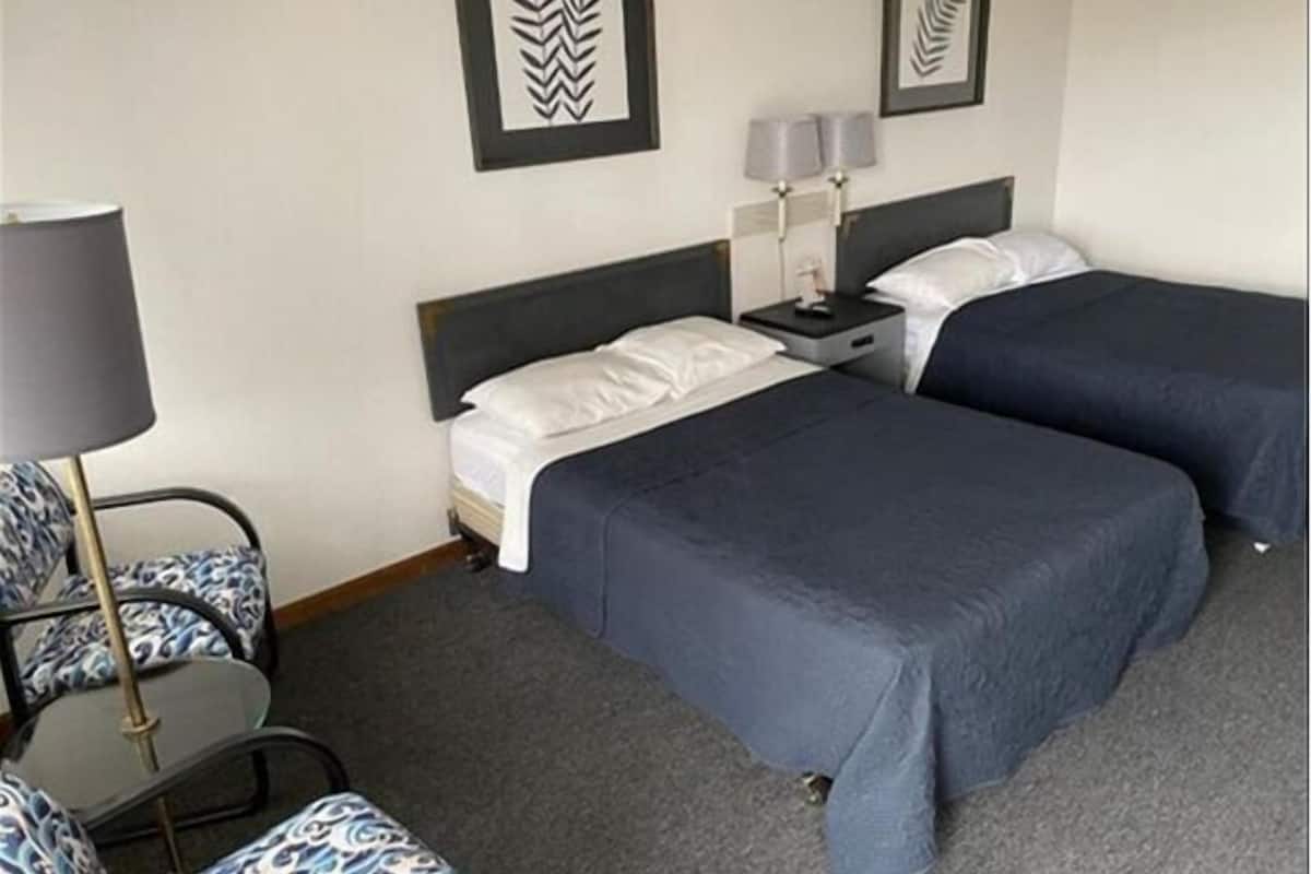A hotel room with two beds, a desk, a chair, and two wall-mounted lamps.