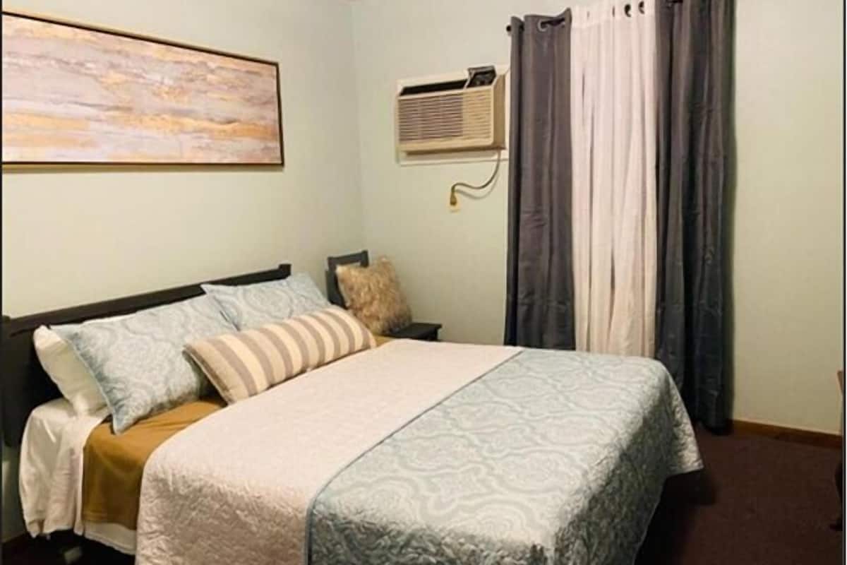 A bedroom with a bed, pillows, a blanket, an air conditioning unit, and curtains.