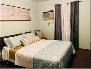 A bedroom with a bed, pillows, a blanket, an air conditioning unit, and curtains. - Rockwood Resort Motel (Lake Ozark)