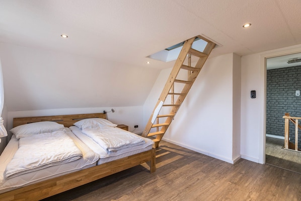 A bedroom with a wooden ladder leading to a skylight, a bed with white bedding, and a wooden floor.