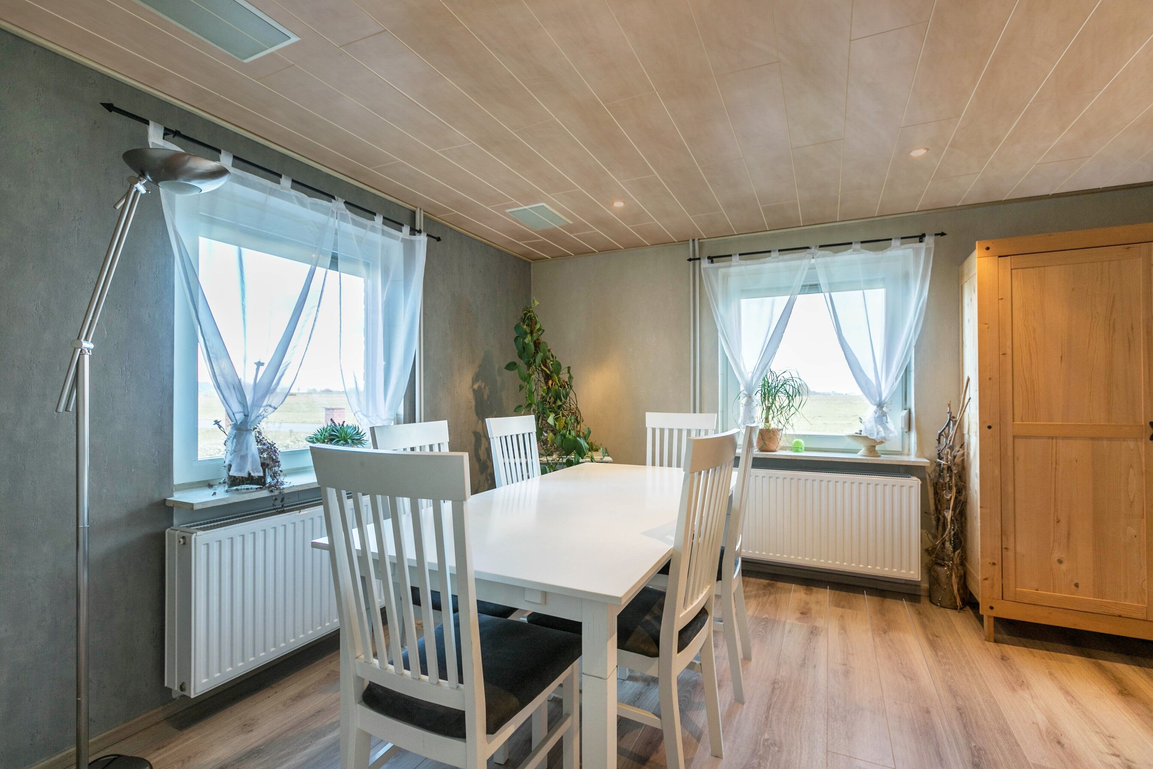 Uma área de jantar com mesa e cadeiras brancas, um abajur e vista através de cortinas leves.