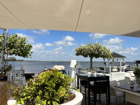 Un espace extérieur aménagé pour les repas, avec vue sur l’eau, comprenant des tables, des chaises et des plantes en pot.