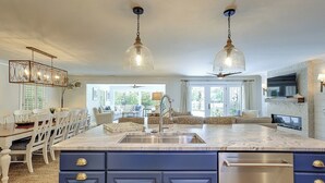 A modern kitchen with a large island, pendant lights, and a dining area with white furniture. - Sunset Rentals | South Live Oak 25 (Hilton Head Island)