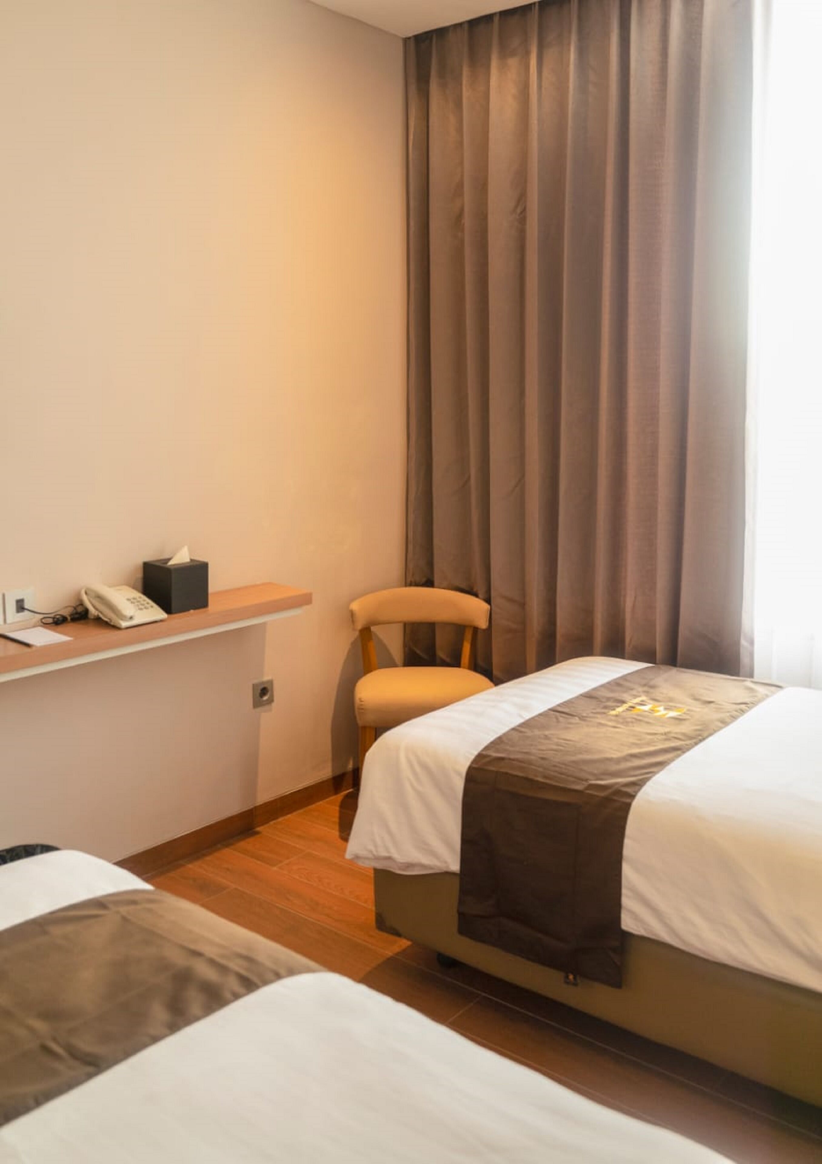 Une chambre d’hôtel avec deux lits, un bureau doté d’un téléphone et d’une boîte de mouchoirs, une chaise et une grande fenêtre avec des rideaux.