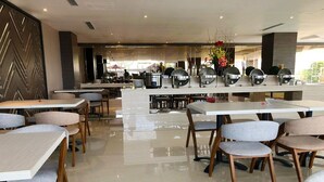 Uma zona de refeições moderna com várias mesas e cadeiras, um balcão central com máquina de café e vista para a área de estar ao ar livre.