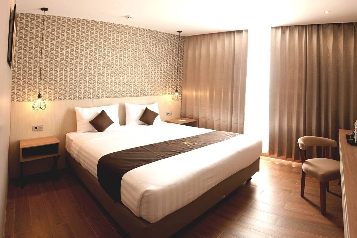 A hotel room with a large bed, wooden flooring, a patterned wall, and a view of the outside through curtains.
