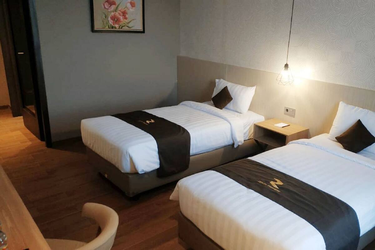 Two single beds with white linens and dark headboards, a wooden floor, a small wooden table, and a wall-mounted light fixture.