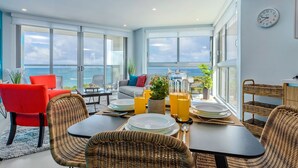 A modern dining area with a view of the sea, featuring wicker chairs, a dining table set with plates and glasses, and a cozy seating area with a sofa and red chairs.
