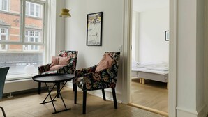 A room with two floral armchairs, a small round table, a bed with white bedding, a framed picture on the wall, and a window with a view of buildings. - City Apartment in Copenhagen with 2 bedrooms sleeps 6 (Copenhagen)
